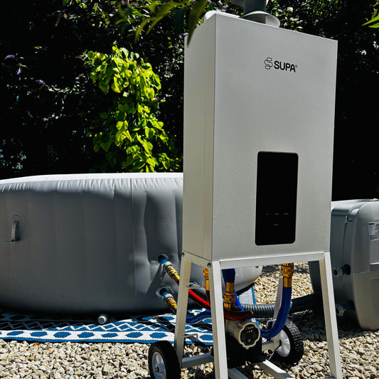 A white portable hot water heater on a wheeled stand, branded SUPA, photographed outdoors on a gravel surface with trees and gray storage tanks in the background.